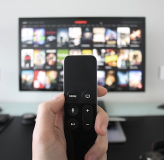 A person holds a remote control in front of a TV displaying a streaming platform interface with various movie and show thumbnails. The room includes a desk with two small lamps on either side of the TV.