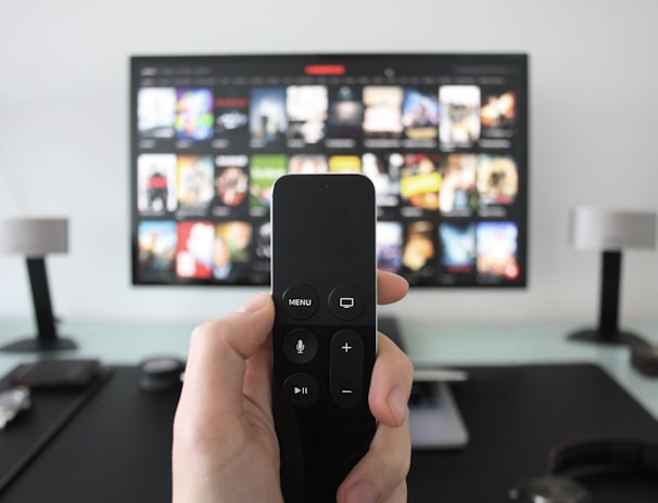 A person holds a remote control in front of a TV displaying a streaming platform interface with various movie and show thumbnails. The room includes a desk with two small lamps on either side of the TV.