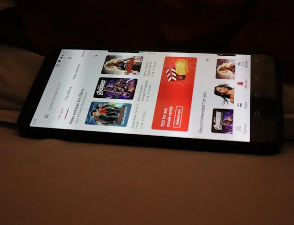 A smartphone displaying a streaming app interface, showing various movie and TV show thumbnails. The screen includes categories like 'For you' and 'New release hit films', with images from popular films. The background is a dimly lit setting with a fabric surface.