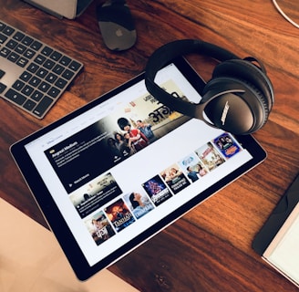 A wooden table holds a tablet displaying a streaming service interface with various movie and TV show thumbnails. Nearby, a pair of Bose headphones rests on the table along with a keyboard and a mouse.