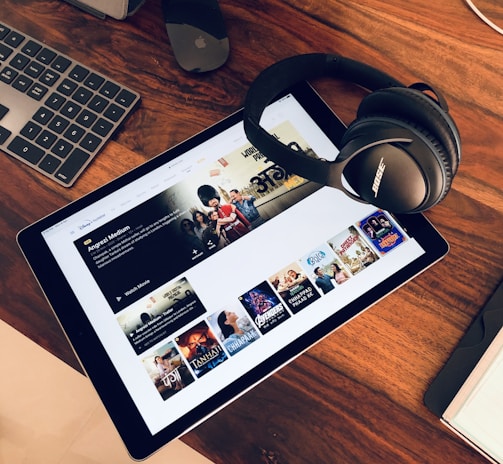 A wooden table holds a tablet displaying a streaming service interface with various movie and TV show thumbnails. Nearby, a pair of Bose headphones rests on the table along with a keyboard and a mouse.