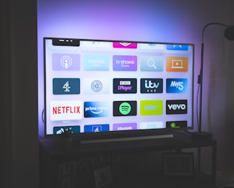 A flat-screen television is displayed in a dimly lit room, showcasing a variety of streaming service apps on its screen, such as Netflix, Prime Video, and Spotify. The TV is positioned on a black stand beside a tall floor lamp, casting a soft ambient light on the surrounding area.