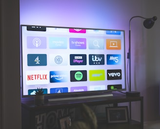 A flat-screen television is displaying a variety of streaming app icons, including Netflix, Prime Video, Spotify, and ITV Hub among others. The TV is placed on a media console with a small potted plant beside it. A tall lamp stands to the right, and a faint, ambient light casts a purple hue on the wall above the TV.