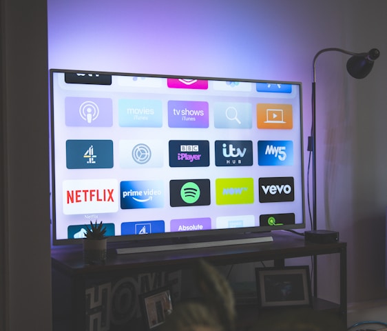 A flat-screen television is displaying a variety of streaming app icons, including Netflix, Prime Video, Spotify, and ITV Hub among others. The TV is placed on a media console with a small potted plant beside it. A tall lamp stands to the right, and a faint, ambient light casts a purple hue on the wall above the TV.