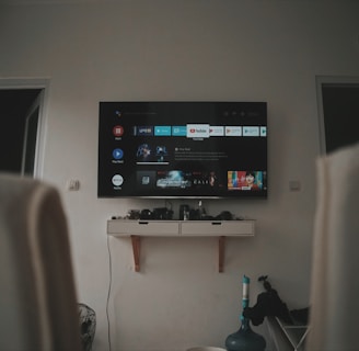 A living room setup featuring a wall-mounted TV displaying a streaming service interface. The room is dimly lit, with the TV as the main light source. Below the TV is a small shelf holding various objects. To the sides, there are partially visible chairs, and in the corner, there is a water bottle on the floor.