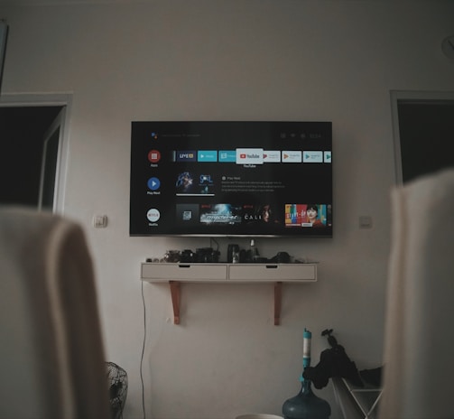 A living room setup featuring a wall-mounted TV displaying a streaming service interface. The room is dimly lit, with the TV as the main light source. Below the TV is a small shelf holding various objects. To the sides, there are partially visible chairs, and in the corner, there is a water bottle on the floor.