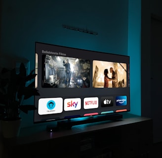 A television screen displaying a user interface with popular streaming service apps including Netflix, Sky, Apple TV, Prime Video, and others. The screen also shows images from movies or shows above the apps. The room is dimly lit with a potted plant visible on the side.