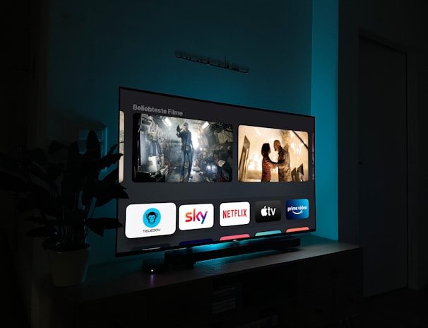 A television screen displaying a user interface with popular streaming service apps including Netflix, Sky, Apple TV, Prime Video, and others. The screen also shows images from movies or shows above the apps. The room is dimly lit with a potted plant visible on the side.