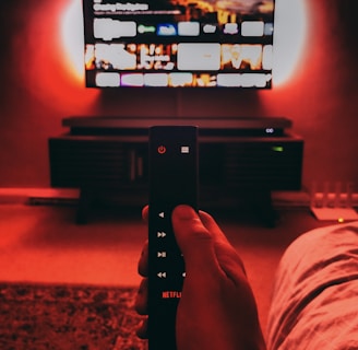 A hand holds a remote with visible buttons, pointed towards a wall-mounted TV displaying a streaming platform interface. The room is dimly lit, with a warm, red ambient light highlighting the TV screen and surroundings.