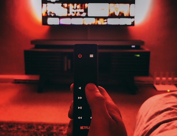 A hand holds a remote with visible buttons, pointed towards a wall-mounted TV displaying a streaming platform interface. The room is dimly lit, with a warm, red ambient light highlighting the TV screen and surroundings.