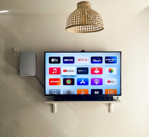A modern living room features a wall-mounted flat-screen television displaying multiple streaming app icons, such as Apple TV, HBO Max, Netflix, Disney+, and Prime Video. Below the TV is a soundbar sitting on a small white shelf. A wicker pendant light hangs from the ceiling above, and the wall is painted a neutral color.