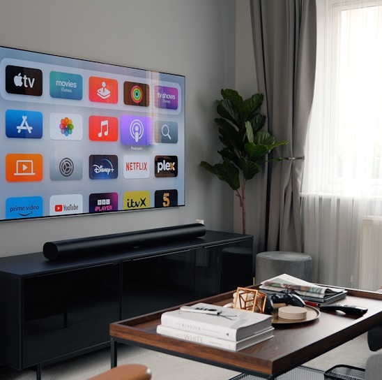 A modern living room featuring a mounted flat-screen TV displaying various streaming service icons. The room includes a black entertainment console beneath the TV, a brown wooden coffee table with books and remote controls, a gray sofa, and a tall green plant near a large window with sheer white curtains.