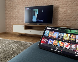 A modern living room features a flat-screen television mounted on a light-colored wooden stand with a patterned wall backdrop. Below the TV, a carpet lies on a wooden floor. In the foreground, a tablet displays a streaming platform interface with various show thumbnails, suggesting a relaxed entertainment setup.