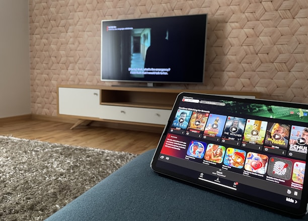 A modern living room features a flat-screen television mounted on a light-colored wooden stand with a patterned wall backdrop. Below the TV, a carpet lies on a wooden floor. In the foreground, a tablet displays a streaming platform interface with various show thumbnails, suggesting a relaxed entertainment setup.